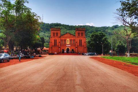 A red brick Catholic cathedral with twin towers stands prominently against a backdrop of lush green hills. The church is approached by a wide red dirt road, with trees lining both sides and a few people and parked cars visible in the foreground.