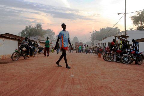 A person in a blue and orange shirt walks across a dusty red dirt street at dusk. Motorcycles are parked along both sides of the road, with small buildings lining the street and other pedestrians visible in the background under a hazy sunset sky.
