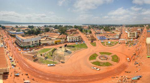 Aerial view of an African city with distinctive red-orange dirt roads, featuring a large roundabout, commercial buildings, and residential areas. A body of water is visible in the background, with the cityscape extending toward distant hills under a partly cloudy blue sky.