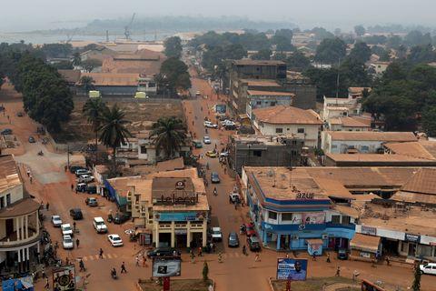 An elevated view of a West African town with reddish dirt roads, commercial buildings with metal roofs, and scattered palm trees. The urban area extends toward a misty body of water in the distance, with various vehicles and pedestrians moving along the main street.