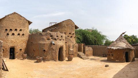 Traditional African mud brick compound featuring multiple adobe structures with distinctive circular ventilation holes. The compound includes larger rectangular buildings with flat roofs, clay pots in the courtyard, and a conical thatched-roof hut on the right. Green trees are visible in the background under a pale blue sky.