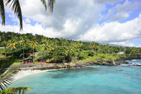 A tropical coastal scene with turquoise blue waters along a rocky shoreline. Palm trees and lush vegetation cover the hillside, with small wooden structures visible near the shore. A small boat floats in the calm bay, and palm fronds frame parts of the image. White clouds float in a blue sky over distant mountains.