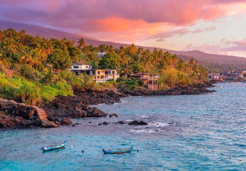  A coastal village at sunset with vibrant pink and orange clouds illuminating the sky. Palm trees and residential buildings dot the rocky shoreline, with traditional outrigger boats floating in the blue-turquoise waters. A volcanic mountain slope rises in the background, covered with tropical vegetation.