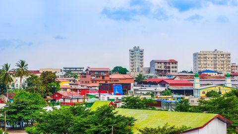 A colorful tropical city skyline with a mix of architectural styles. Various buildings with bright red, blue, yellow, and green roofs are visible among palm trees and vegetation. Several taller apartment buildings rise in the background under a clear blue sky with scattered clouds.