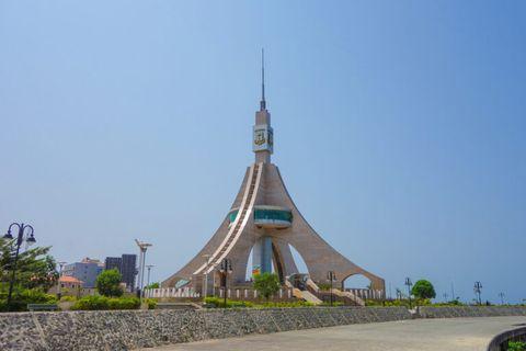 A striking modern monument or memorial structure with a distinctive conical shape and tall spire rising from the center. The building features curved concrete elements that sweep upward, with a band of teal-colored windows around its middle section. The structure sits on landscaped grounds with a clear blue sky in the background.