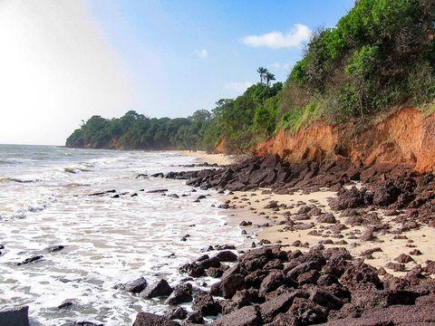 A coastal beach with dark volcanic rocks scattered across golden sand, where waves wash onto the shore. Red-orange clay cliffs rise above the beach, topped with dense tropical vegetation and palm trees. The coastline curves into the distance under a clear blue sky, creating a striking contrast between the dark rocks, sandy beach, reddish cliffs, and green foliage.