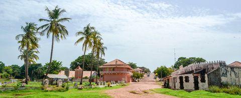 A small village road in a tropical setting with tall palm trees lining the path. A distinctive pink colonial-style building with a balcony stands in the center, while older weathered structures are visible on the right. The unpaved dirt road extends into the distance under a bright cloudy sky. Some gravestones or markers are visible in a grassy area on the left.