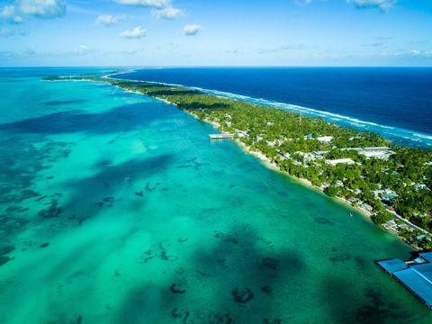 Aerial view of a narrow tropical atoll with turquoise lagoon water on one side and deep blue ocean on the other, lined with palm trees and small buildings along its shore.