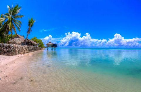  Idyllic tropical beach with traditional thatched-roof huts including one on stilts over shallow, transparent turquoise water, palm trees, and white sand against a vibrant blue sky with fluffy clouds.