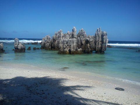 Jagged limestone rock formations rising from clear turquoise waters at a tropical beach with white sand and gentle waves breaking in the distance under a clear blue sky.