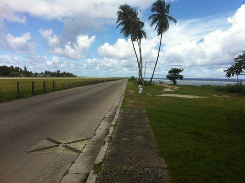 Coastal road running alongside the ocean with tall palm trees in the center, grassy areas on both sides, a fence line to the left, and the ocean visible to the right under a partly cloudy sky.