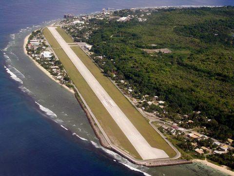 Aerial view of a narrow island runway stretching along the coastline with ocean waves breaking along the shore, dense green vegetation covering most of the island, and small settlements visible along the coastline.