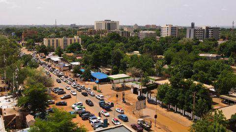 Aerial view of an African city with busy streets filled with cars and traffic, surrounded by lush green trees and vegetation, with multi-story buildings and high-rises visible in the background under a hazy sky.
