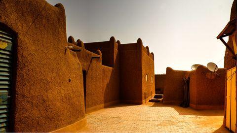 Traditional mud-brick architecture featuring thick earthen walls with rounded corners and distinctive crenellated rooflines creating a courtyard space with a paved floor. Satellite dishes are mounted on one wall, and the warm brown structures are illuminated by golden sunlight against a bright sky.