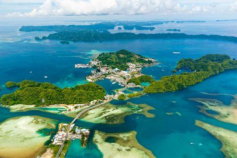 Aerial view of a Pacific island nation showing a settlement connected by bridges or causeways, surrounded by vibrant turquoise waters, coral reefs, and numerous green forested islands stretching to the horizon under a partly cloudy sky.