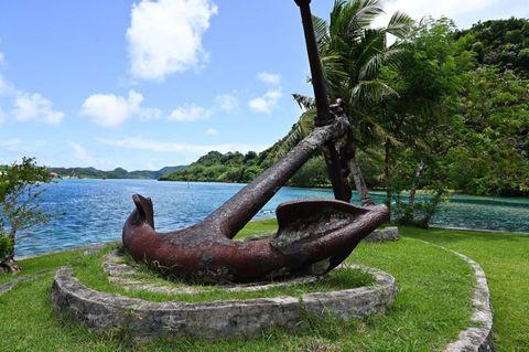 Large rusty ship anchor displayed as a monument on a circular stone platform on a grassy shoreline, with calm blue water and lush tropical vegetation in the background under a bright blue sky with scattered clouds.
