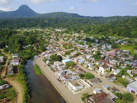 Alt text: Aerial view of a small coastal town nestled between lush green mountains and a river. Houses with white and colored roofs are arranged along streets, with dense tropical forest covering the surrounding hills and a prominent mountain peak in the background.
