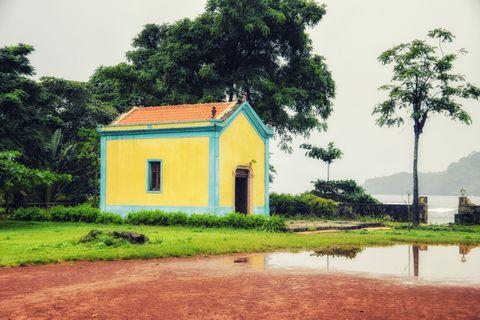 A charming yellow building with turquoise blue trim and an orange tile roof, surrounded by lush green vegetation. The small structure stands near a body of water with reflective puddles on reddish soil in the foreground and tall trees providing shade.