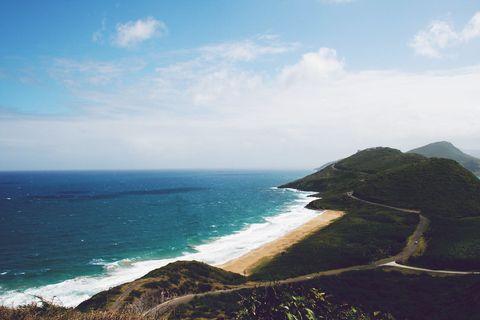 Coastal landscape featuring a curving shoreline with golden sand beach and turquoise waters. A winding road follows the green hillside contours with mountains visible in the distance under a partly cloudy sky.