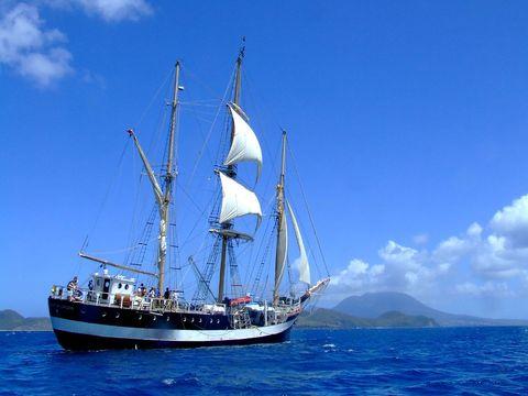  Traditional sailing ship with white sails unfurled against a brilliant blue sky. The three-masted vessel carries passengers on the deep blue ocean with volcanic islands visible in the background.