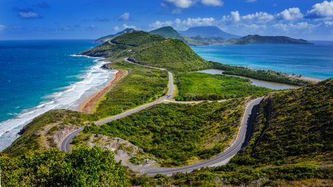 Aerial view of a coastal road winding along a lush green peninsula with turquoise water on both sides. The scenic highway curves through tropical vegetation with mountains in the background under a bright blue sky with fluffy white clouds.