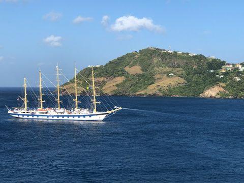 A majestic tall sailing ship with golden masts and white sails anchored in the deep blue waters near a lush green island with scattered hillside homes under a clear blue sky.
