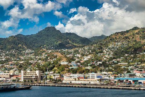 A scenic view of Kingstown, Saint Vincent and the Grenadines, with colorful hillside houses, lush green mountains, a busy waterfront, and a vibrant sky with clouds.