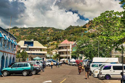 A bustling street scene in Kingstown, Saint Vincent and the Grenadines, with parked cars, pedestrians, and market activity, set against a lush green hillside with colorful houses and dramatic clouds in the background.