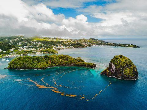 Aerial view of a lush green island and a rugged rock formation surrounded by vibrant blue ocean waters, with a coastal town in the background under a partly cloudy sky.