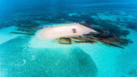 Aerial view of a small sandy island surrounded by crystal-clear turquoise waters, with a single thatched umbrella in the center, nestled among coral reefs.