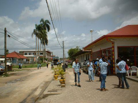 A street scene in what appears to be a tropical town with palm trees, showing people in light blue uniforms walking on a paved sidewalk next to a dirt road. Buildings with red roofs line the street, and utility poles with power lines are visible overhead under a partly cloudy sky.