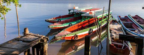 Colorful wooden fishing boats in red, green, and white tied to a simple wooden dock on calm reflective water. The boats are traditional in style and appear to be used by local fishermen.