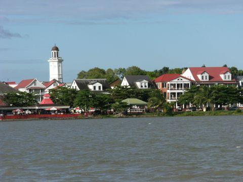 A waterfront view of a colonial-style town with white and red-roofed buildings, featuring a prominent white lighthouse or clock tower. The buildings are arranged along the shoreline with trees interspersed between structures, viewed from water that appears brownish in color.
